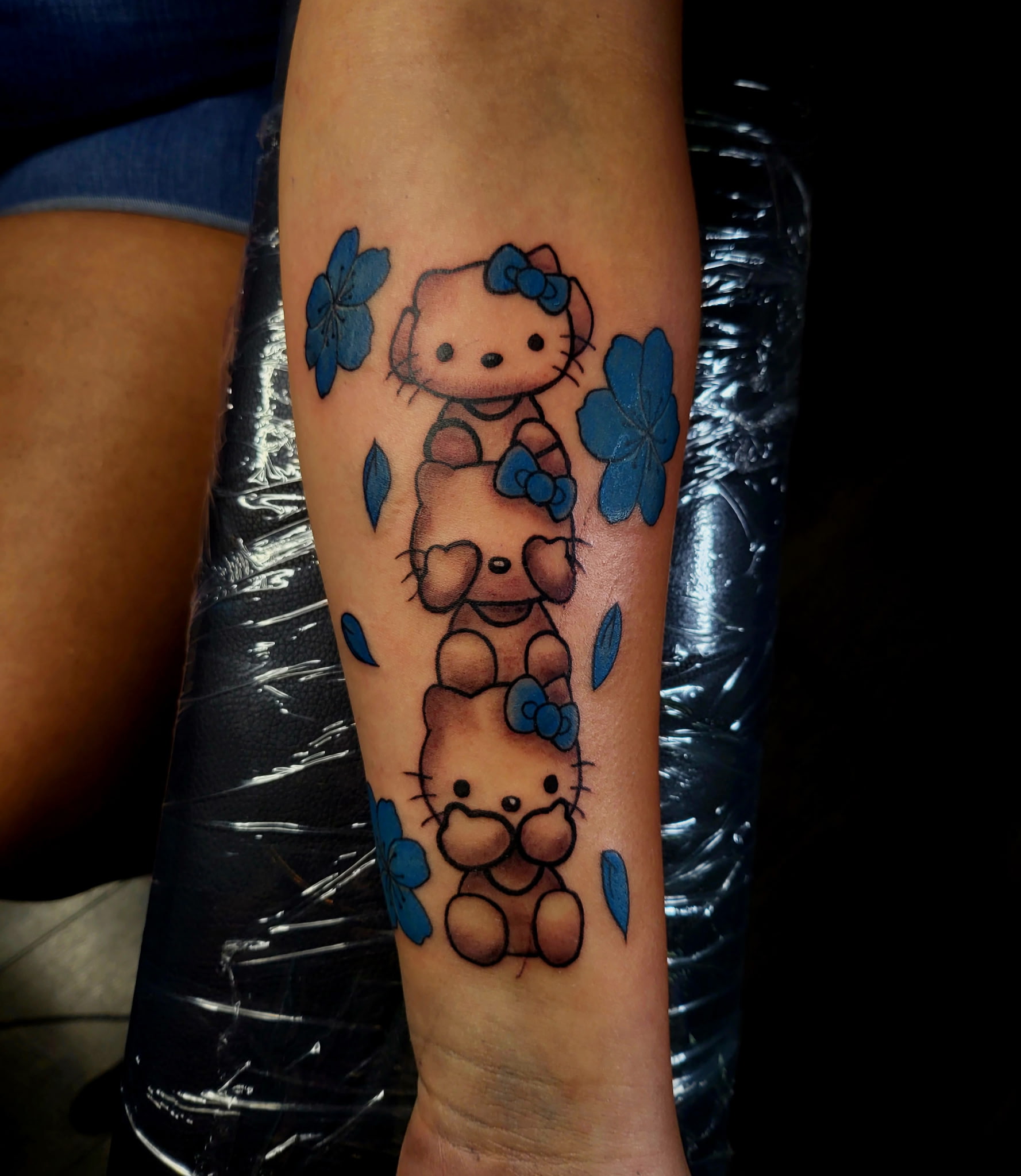 Forearm tattoo of three stacked cartoon cats with blue cherry blossoms