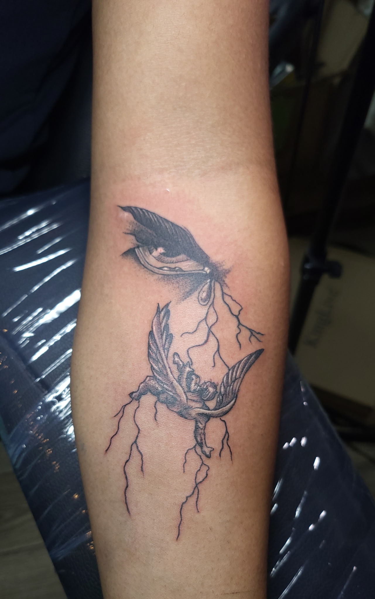 Forearm tattoo of an eye with a falling winged figure and lightning